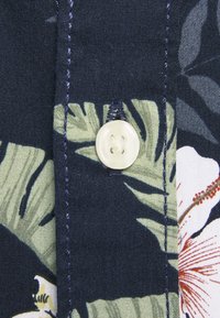 Short-sleeve shirt featuring a dark navy background with green tropical leaf patterns and a cream button, highlighted by contrasting stitching.