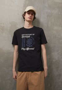Zwarte katoenen T-shirt met meerkleurige tekstgraphics, korte mouwen en een relaxte pasvorm. Gecombineerd met beige shorts voor een neutrale achtergrond.