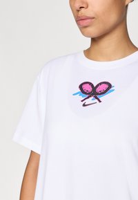 Λευκό μπλουζάκι με κοντό μανίκι που διαθέτει ένα ροζ και μαύρο γραφ ικό τένις στο κέντρο. Στο λαιμό στρογγυλό, μαλακή υφή.