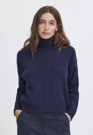 Femme aux cheveux bruns ondulés portant des boucles d'oreilles argentées en créoles, un pull à col roulé côtelé bleu marine et un pantalon bleu marine avec les mains dans les poches.