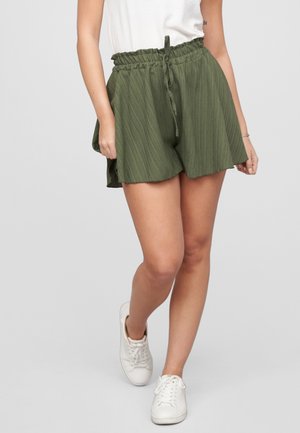 Vrouw draagt olijfgroene shorts met hoge taille en trekkoord, een wit topje en witte sneakers, lopend tegen een effen achtergrond.