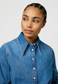 Lyseblå denimskjorte med spiss krage, trykknapper og buet bærestykke. Stoffet har en teksturert overflate og et uformelt design.