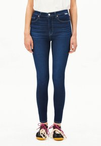 Dunkelblaue, hoch taillierte Skinny-Jeans mit fünf Taschen, einem Knopfverschluss und hellen Nähten, kombiniert mit schwarzen Sneakers.