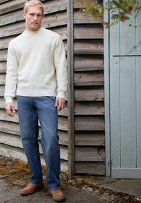 Pull en maille torsadée crème, jeans en denim bleu et chaussures en daim marron ; devant un mur en bois vieilli et une porte grise.