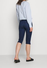 Tummansinisiä farkku capri-housuja, joissa on muotoiltu malli, lyhyt pituus ja takataskut. Yhdistetty vaaleansiniseen pitkähihaiseen paitaan.