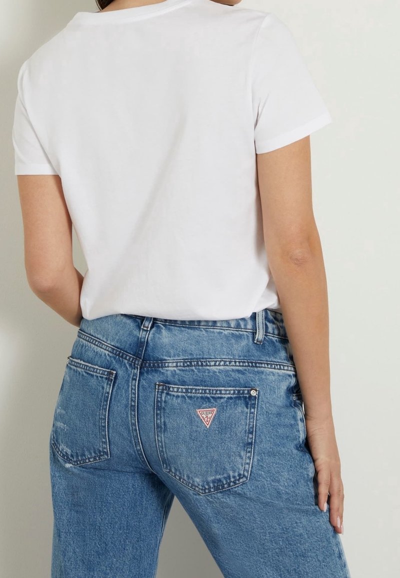 Personne portant un t-shirt blanc uni à manches courtes et un jean bleu avec un logo triangulaire sur la poche arrière droite, debout de côté.