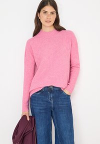 Różowy sweter wykonany na drutach z wysokim kołnierzem i prążkowanymi mankietami, w zestawie z niebieskimi dżinsami. W lewej ręce trzymany jest bordowy przedmiot.