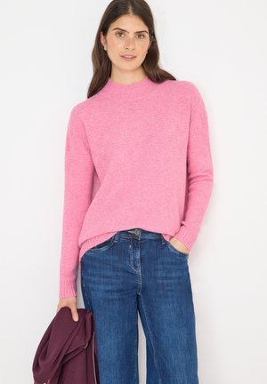 Lyserød striktrøje med høj hals og ribbede manchetter, sammen med blå denimjeans. En bordeaux genstand holdes i venstre hånd.