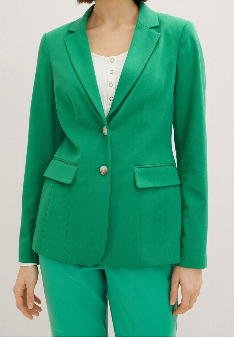 Femme portant un blazer vert cintré à deux boutons sur un haut blanc, assorti à un pantalon vert, se tenant devant un fond uni.