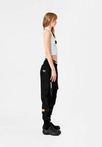 Pantalones cargo negros con múltiples bolsillos y cremalleras, combinados con un top corto blanco. Ella lleva altavoces negros texturizados, diseñados para el confort.