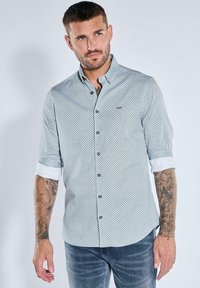 Hellblau gemustertes Hemd mit Knöpfen und Kragen, hochgesteckten Ärmeln und dunklen Knöpfen, kombiniert mit blauen Jeans. Verfügt über ein dezentes Logodetail.