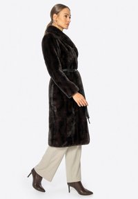 Bruine nepbont jas met een sjawl kraag, ceintuur in de taille en lange mouwen; gepresenteerd met beige broeken en bruin haklaarsjes.