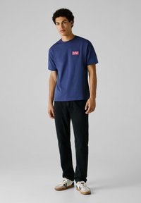 Donkerblauwe katoenen T-shirt met een ronde hals en korte mouwen, voorzien van een rood en wit logo-opdruk op de linkerborst. Zwarte jeans en sneakers.