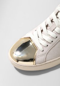 Baskets en daim beige clair avec lacets blancs et embout métallique doré réfléchissant sur un fond gris clair.