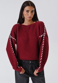 Donna con lunghi capelli scuri che indossa un maglione rosso lavorato a maglia con dettagli in filato bianco attorcigliato sulle maniche e sulle spalle, abbinato a jeans neri.
