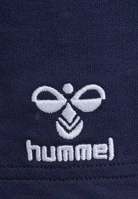 Marinblått tyg med vitt broderat logo med abstrakt design och texten "hummel." Texten är placerad under logotypen med strukturerad sömnad.
