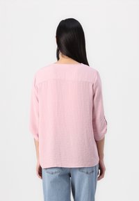 Blusa rosa chiaro, texture, con maniche a tre quarti, taglio dritto e scollatura rotonda. Dettagli delle cuciture visibili lungo il retro.