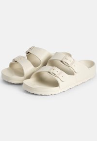Cream-colored sandals with two adjustable straps, made of textured rubber material, featuring a contoured footbed and a durable sole.