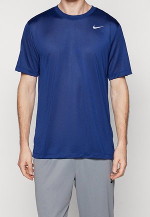 Homme portant un t-shirt de sport Nike bleu uni à manches courtes et un short gris clair, debout devant un fond uni.