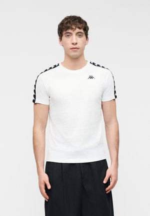 Ung mand iført en hvid t-shirt med sorte logo-striber på skuldrene og sorte bukser, stående foran en ensfarvet baggrund.