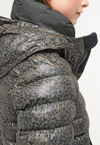 Kurtka ocieplana z wysokim kołnierzem, wykonana z szarej tkaniny z geometrycznym, ciemnym wzorem. Zawiera zamki błyskawiczne i detale przeszyć.