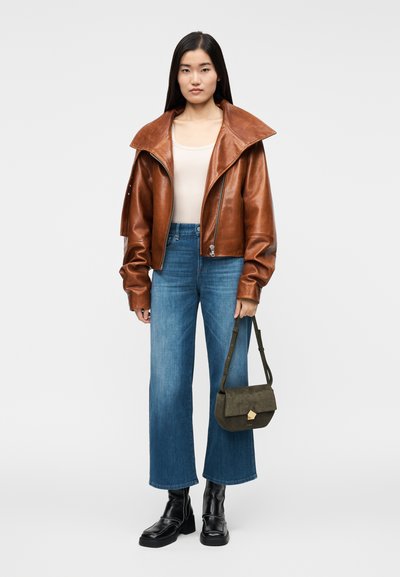 Giacca corta in pelle marrone con colletto largo, canottiera chiara, jeans a vita alta blu, stivaletti neri alla caviglia e borsa verde oliva con dettaglio geometrico.