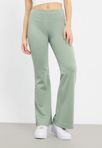 Pants de sport vert clair évasés en tissu doux avec une taille haute, portés avec des baskets blanches ; texture lisse et design ajusté.