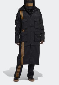 Černý dlouhý kabát s mnoha kapsami, zvýrazněný zlatými pruhy po stranách. Tkanina vypadá matně, má strukturovaný design a kapuci.