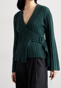 Top verde con tejido acanalado, con un escote en V profundo y una cinturilla lateral. Mangas largas con puños acampanados y tela texturizada para mayor profundidad visual.