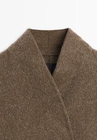 Collo in morbido e peloso tessuto di lana marrone chiaro con ampi rever su sfondo bianco, che mostra la texture del tessuto e i dettagli delle cuciture.