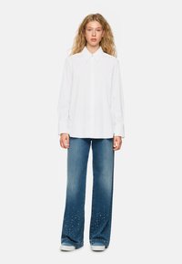 White collared shirt, long sleeves, paired with blue wide-leg jeans featuring studded accents at the hems. Model stands centrally.
