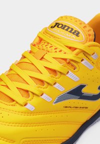 Φωτεινό κίτρινο αθλητικό παπούτσι Joma Maxima με κίτρινες κορδόνια, επάνω μέρος από δίχτυ, μαύρο λογότυπο και λεπτομέρειες συστήματος αερισμού στο πλάι.