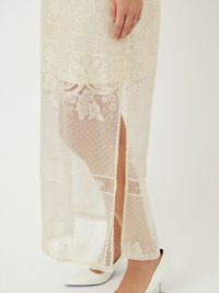 Robe en dentelle ivoire avec des motifs floraux complexes, en tissu transparent et dotée d'une fente latérale. Associée à des escarpins blancs à bout pointu.