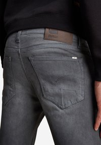 Jeans de mezclilla gris con un parche de cuero etiquetado "RAW" en la cintura, con bolsillos traseros estándar y detalles de costura sutiles.