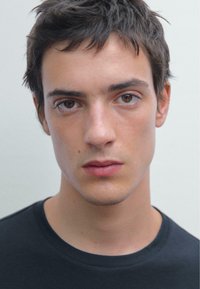 Detailní záběr na mužského modela s krátkými tmavými vlasy a světlou pletí, který nosí jednoduché černé tričko na neutrálním pozadí.