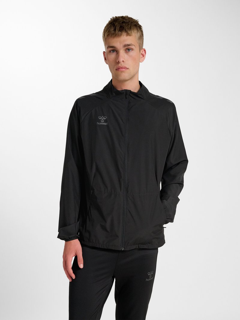 Junger Mann trägt eine schwarze Sportjacke mit Reißverschluss und passende Hose, steht mit einer Hand in der Tasche vor einem schlichten weißen Hintergrund.