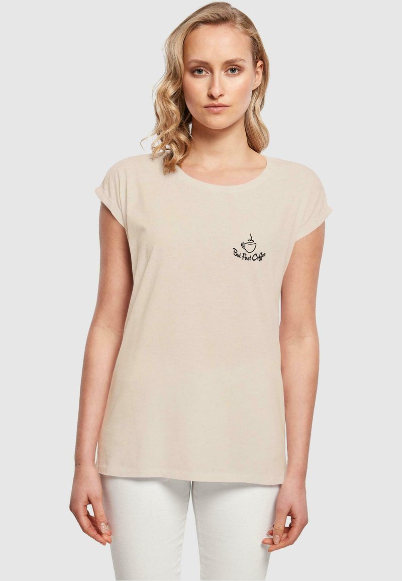 Jasno beżowy t-shirt wykonany z miękkiego materiału, z krótkimi rękawami i zaokrąglonym dekoltem; na przodzie znajduje się logo czarnej filiżanki kawy.
