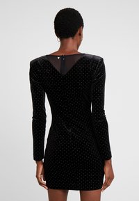 Schwarzes Samtkleid mit langen Ärmeln, versehen mit weißen Punkten, einem V-Ausschnitt hinten und einem verdeckten Reißverschluss am Hals. Glatte Textur.