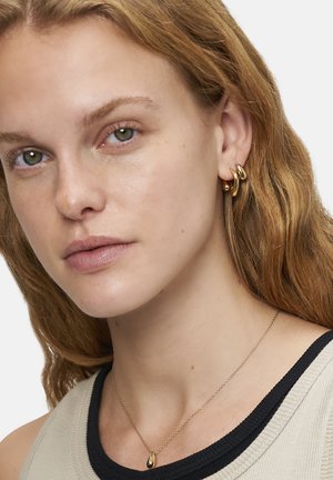 Goldene Creolen in verschiedenen Formen und eine Tropfenanhänger-Halskette. Das Model hat wellige braune Haare und trägt ein schwarzes und beiges Oberteil.