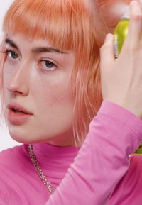 Jeune personne avec des cheveux courts couleur pêche qui vaporise de la laque sur le côté tout en portant un haut à manches longues rose et un collier en chaîne argentée.