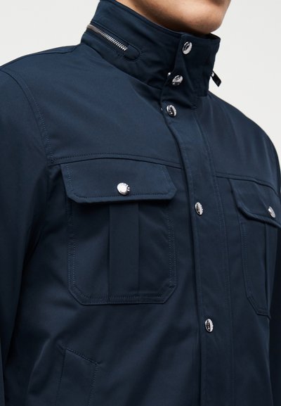 Άνδρας που φοράει μπλε σκούρο μπουφάν με ψηλό γιακά, δύο τσεπάκια στο στήθος με κουμπιά, κάθετες ραφές και ασημένια κουμπωτά κουμπιά μπροστά.