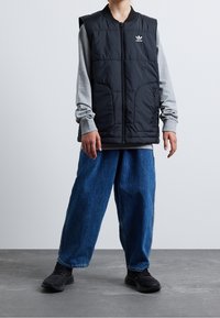 Gilet imbottito nero con chiusura a zip e due tasche frontali, indossato sopra una maglietta grigia a maniche lunghe, abbinato a jeans blu larghi e scarpe nere.