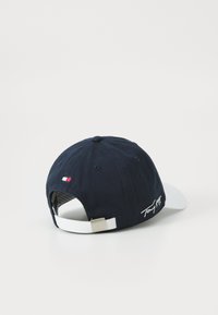 Navy baseball cap with a white visor, featuring the brand logo embroidered in white and a small tricolor flag patch on the back strap.
