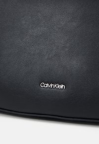 Černá kožená kabelka s texturovaným povrchem a stříbrným akcentem loga "Calvin Klein". Hladké okraje a minimalistický design.