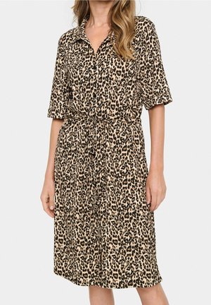 Femme portant une robe léopard longueur genou avec manches courtes et ceinture nouée à la taille, debout devant un fond blanc uni.
