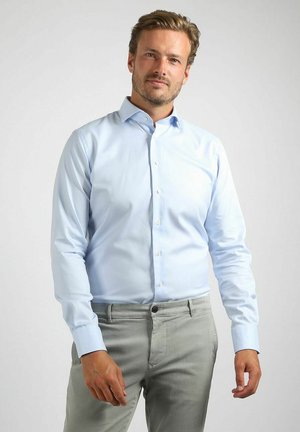 Man staat en draagt een lichtblauw overhemd met knopen en een lange mouw, samen met lichtgrijze broek, tegen een effen achtergrond.