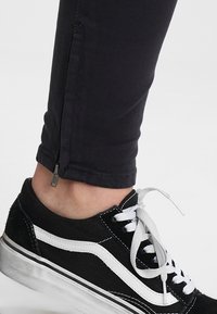 Black slim-fit pants feature a zippered ankle detail, paired with black and white canvas sneakers with white laces and side stripe.