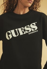 Zwarte sweatshirt met geribbelde halslijn, voorzien van een gedurfde witte "GUESS ORIGINALS" logo in een verouderde stijl over de voorkant.