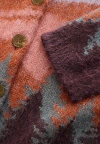 Puha textúrájú kötött pulóver, multicolor cikkcakk mintával narancs, rózsaszín, szürke és sötétlila színekben, nagy barna gombokkal díszítve.