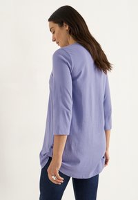 Kvinna klädd i en lång lavendelfärgad topp med trekvartsärmar och mörka jeans, ståendes i profil mot en enkel ljus bakgrund.